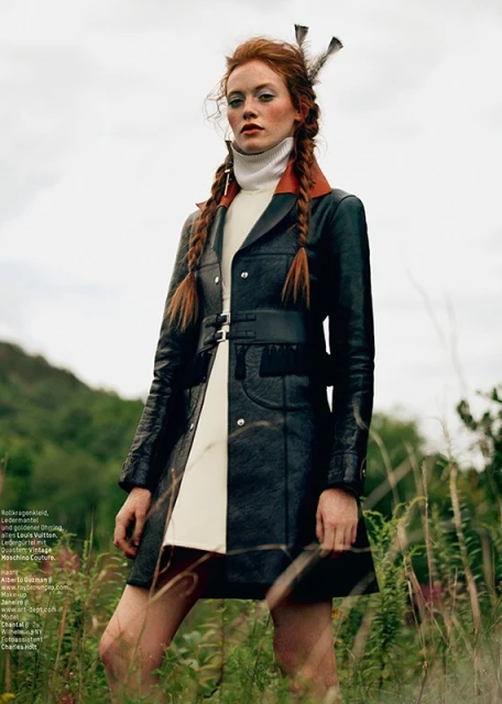 A model with red hair in pigtails poses outdoors, wearing a black leather coat, white turtleneck, and feathers in her hair, in a fashion editorial.