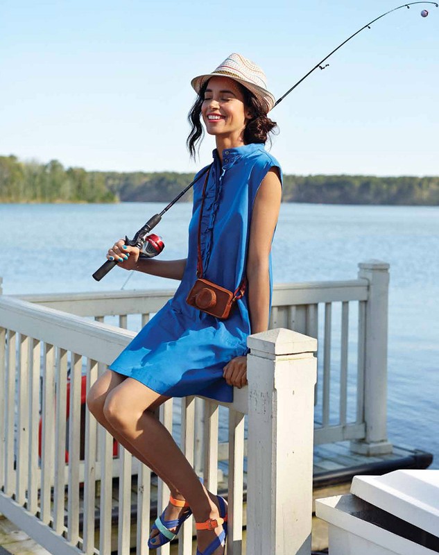 Woman in blue dress and hat sits on railing with fishing rod by lake, enjoying outdoor leisure on a bright day.