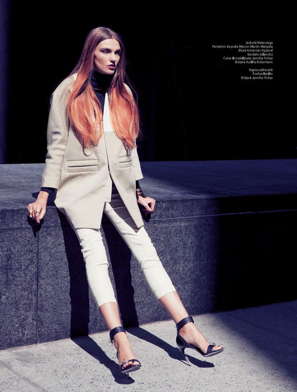 A slender fashion model poses outdoors sitting on a stone ledge, wearing a beige coat, black top, white pants and high heels.
