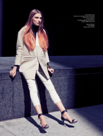 A slender fashion model poses outdoors sitting on a stone ledge, wearing a beige coat, black top, white pants and high heels.