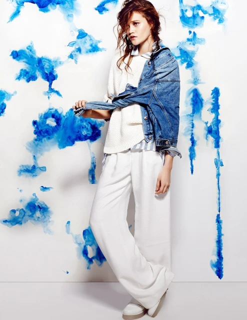 Fashion shot of a young woman in a collared white sweater and white pants standing in front of a white and blue watercolor-painted wall.
