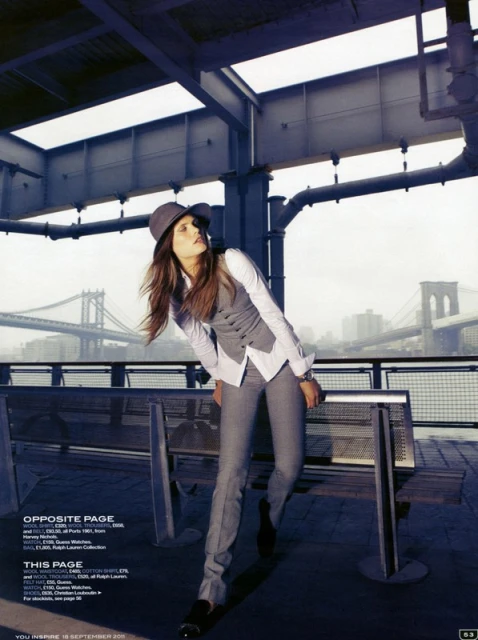 Fashion shot of an adult female in a gray pantsuit and hat posing outside on a city overlook with a bridge and cityscape in the background.