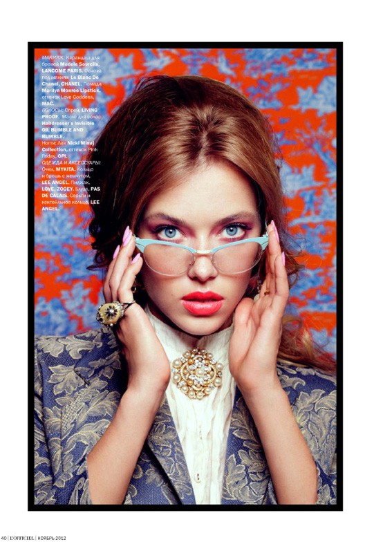 Fashion portrait of a young adult woman with blue eyes wearing glasses, a jacket, and a brooch in an indoor studio setting.