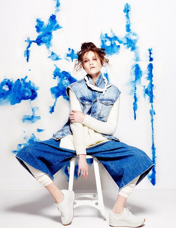 A woman in wide-legged jeans and denim vest sits on a white stool in a studio with blue painted splashes on the white background.