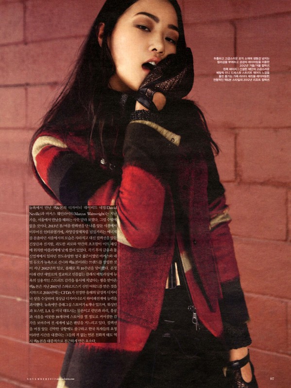 Close-up portrait of a woman wearing a red and black patterned jacket and dark gloves against a red brick wall