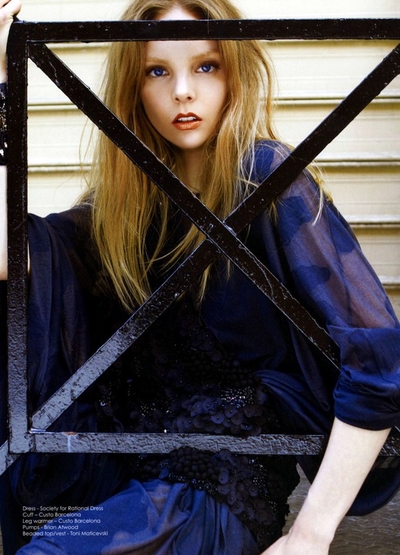 Blonde woman in sheer dark dress poses indoors behind a dark metal frame, creating a bold fashion image with natural light.