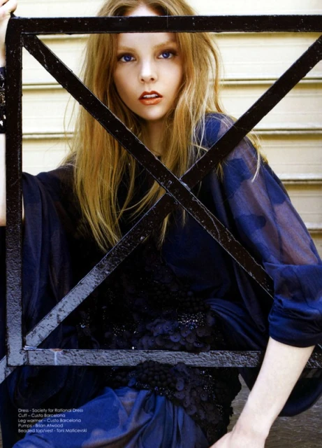 Blonde woman in sheer dark dress poses indoors behind a dark metal frame, creating a bold fashion image with natural light.