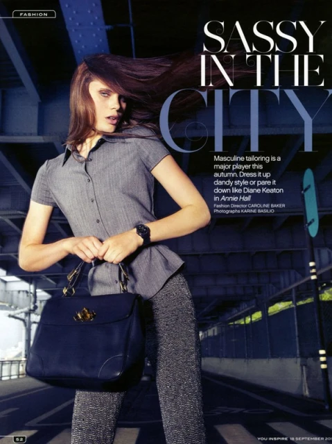 Fashion editorial featuring a woman in gray button-down shirt and patterned pants, holding a black handbag under a concrete structure.