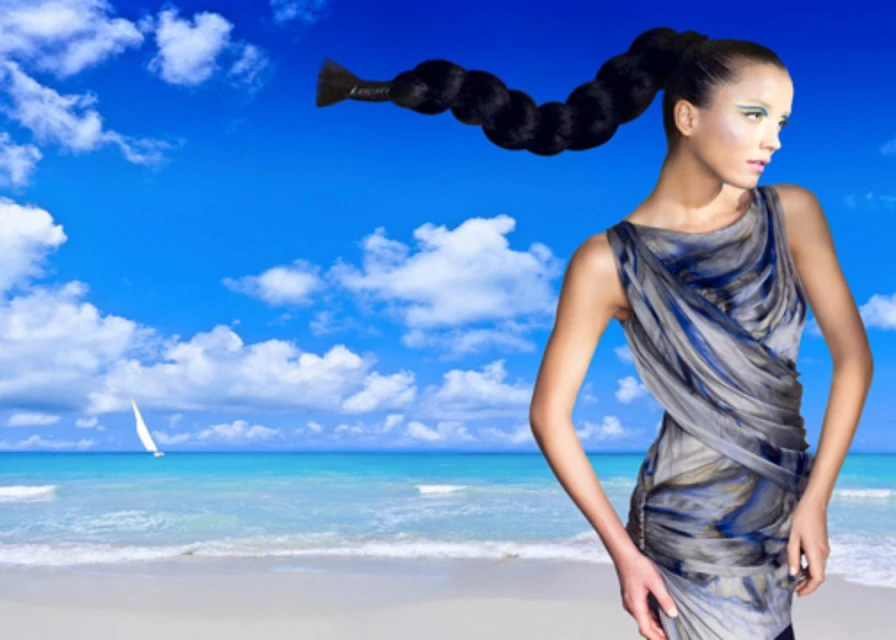 Woman in a draped dress on a beach with turquoise ocean, blue sky, and white sailboat. Long braided hair extends across the scene.