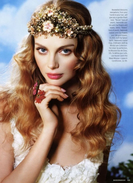 Close-up of a fair-skinned model with reddish-blonde wavy hair, wearing a floral headpiece and ring, against a light blue sky backdrop.
