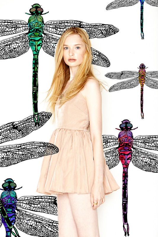 Young woman in a sleeveless dress posing in front of a white backdrop with illustrations of colourful dragonflies and their wings.