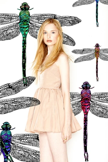 Young woman in a sleeveless dress posing in front of a white backdrop with illustrations of colourful dragonflies and their wings.