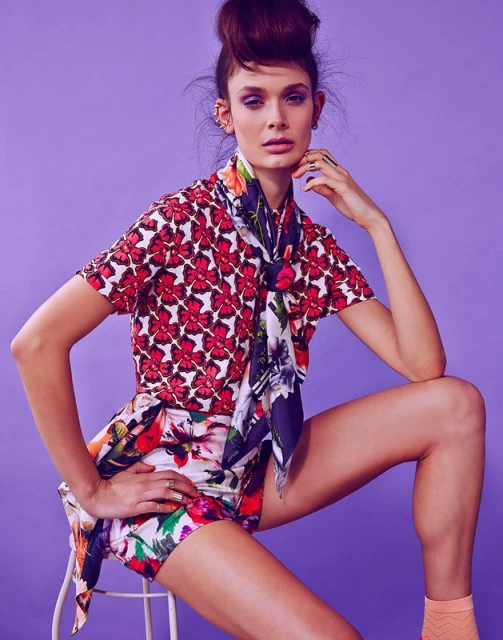 Fashionable woman poses in a floral-patterned ensemble with a matching scarf, seated against a lilac background in a studio setting.