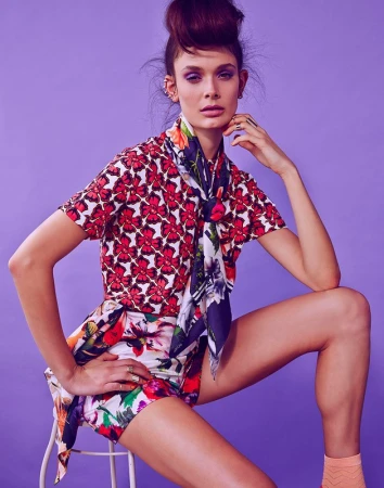 Fashionable woman poses in a floral-patterned ensemble with a matching scarf, seated against a lilac background in a studio setting.