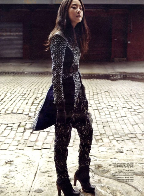 A young woman poses in an urban alleyway. She wears an intricately patterned black and gray dress, long black boots, and black gloves.