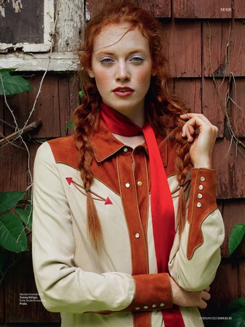 Red-haired woman wearing a Western-style shirt and a red scarf posing outdoors with weathered wood siding in the background.