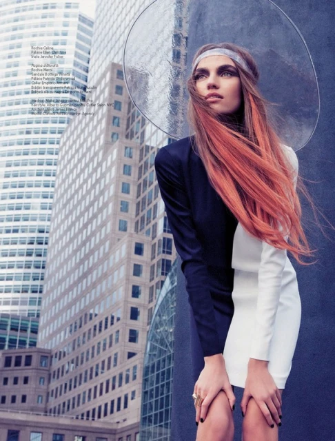 Fashion editorial shot of a young adult female model with red-dyed hair wearing a color-blocked dress with tall office buildings in the background.