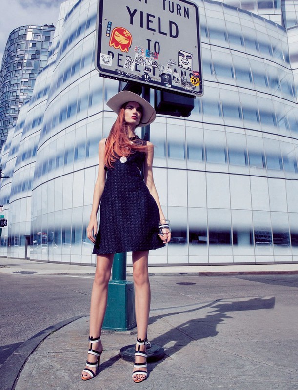 A red-haired woman in a dark dress and light hat stands near a sign in an urban environment with a modern glass building behind her.