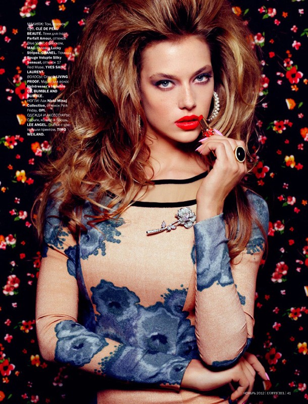 Fashion editorial image: A blonde woman applies red lipstick against a dark floral background. She wears a floral print dress.