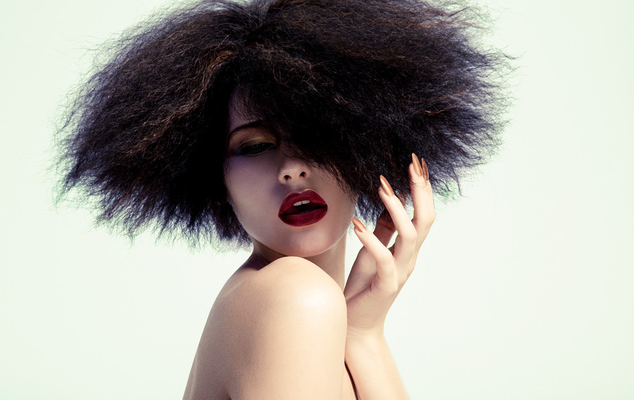 Glamorous woman with dark, voluminous Afro hairstyle, bold red lipstick, and golden nails poses against a soft, light-green background.
