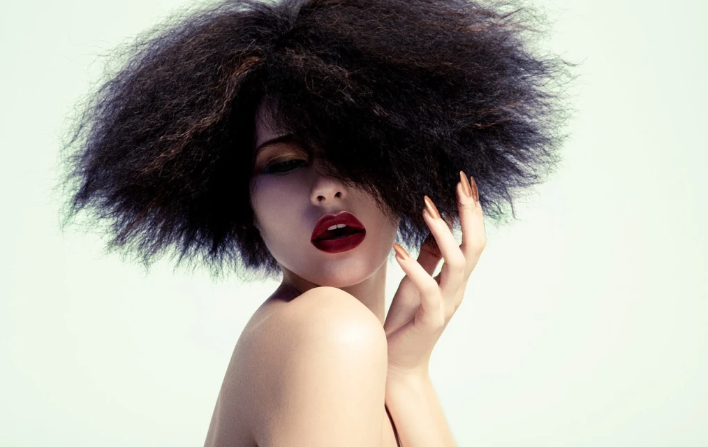 Glamorous woman with dark, voluminous Afro hairstyle, bold red lipstick, and golden nails poses against a soft, light-green background.