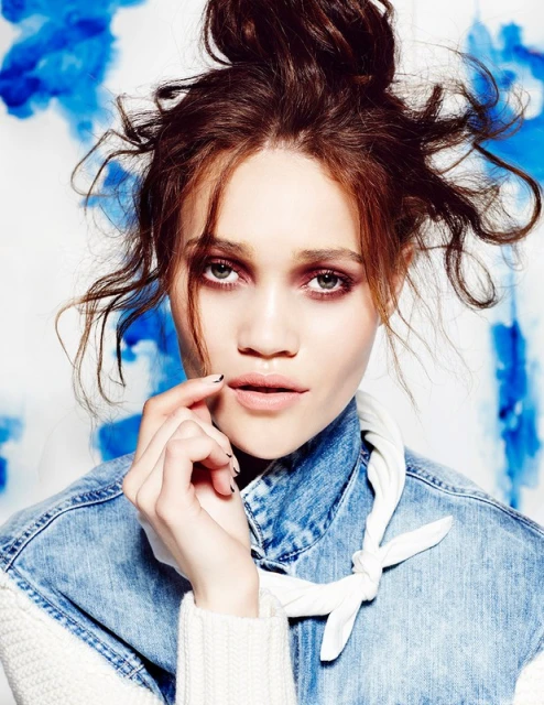 Close-up portrait of a young adult female with a messy bun hairstyle wearing a denim jacket, set against a white and blue background.
