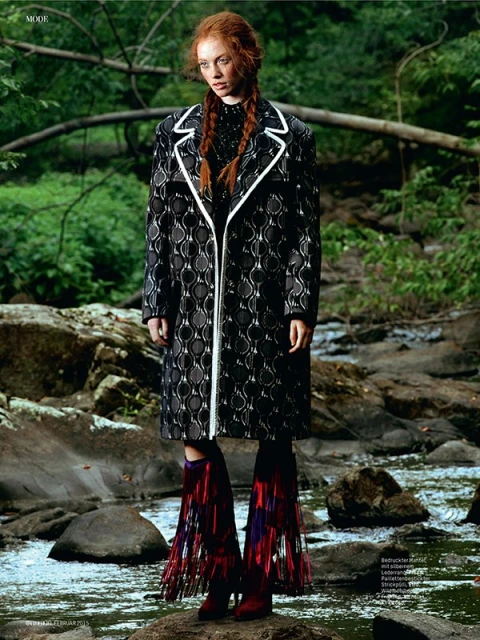 Fashion editorial shot of a red-haired woman in a black and white coat and fringed boots standing on rocks in a shallow creek.