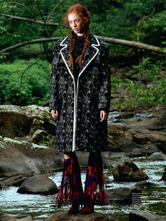 Fashion editorial shot of a red-haired woman in a black and white coat and fringed boots standing on rocks in a shallow creek.