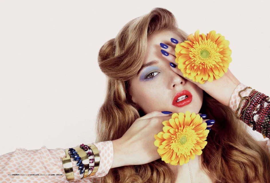 A young woman poses in a studio setting with yellow Gerbera daisies covering her eyes, wearing blue nail polish and bold makeup.