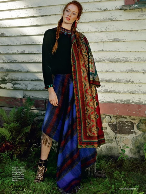 Fashionable woman in long-sleeve top and blue plaid wrap poses outdoors by rustic weathered building with green foliage; editorial.