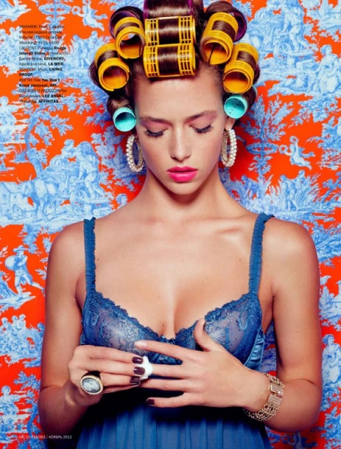 Portrait of a woman with hair curlers wearing a blue dress and jewelry against an orange patterned backdrop in a beauty-style fashion shot.