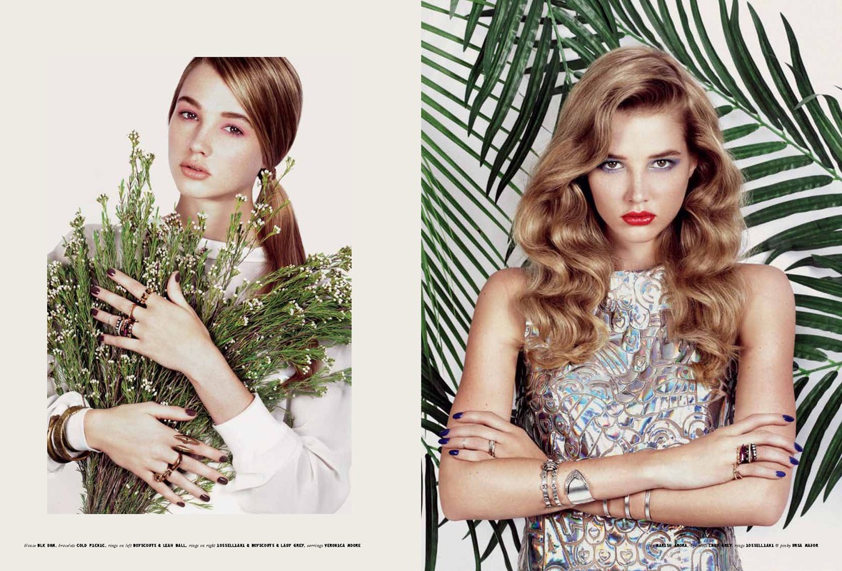 Editorial fashion shot of two young adult women wearing jewelry and makeup. One holds flowers, the other poses with tropical leaves.