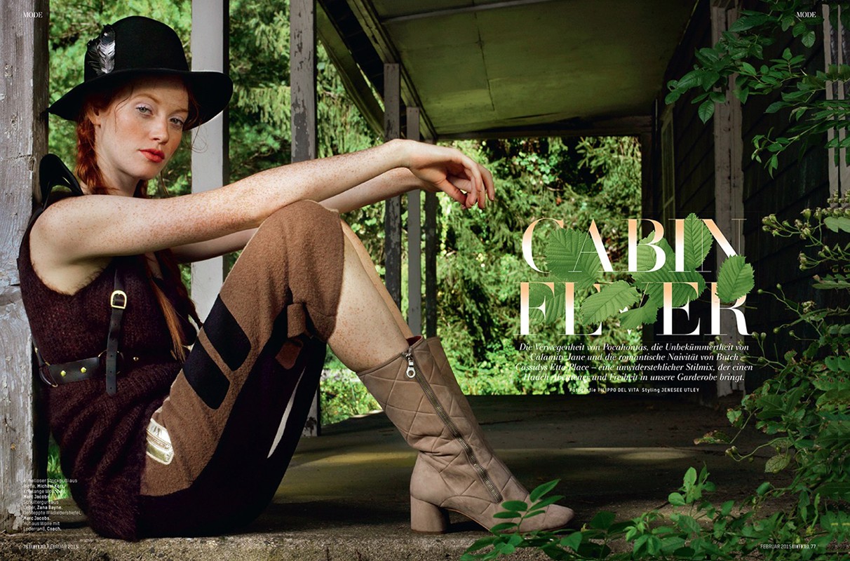 Fashion editorial shot of a red-haired woman in a sleeveless top, patterned pants, and boots, sitting inside a cabin with a forest backdrop.