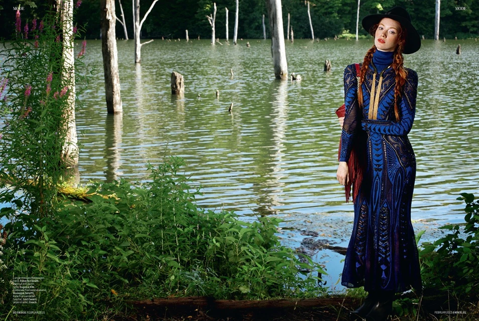 A red-haired woman in a dark blue dress and black hat stands near a lake surrounded by trees and greenery in an outdoor fashion editorial.