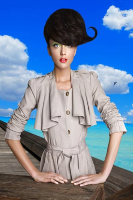 A woman poses in a beige outfit with an elaborate hairstyle against a coastal backdrop with a wooden pier, blue water, and cloudy skies.
