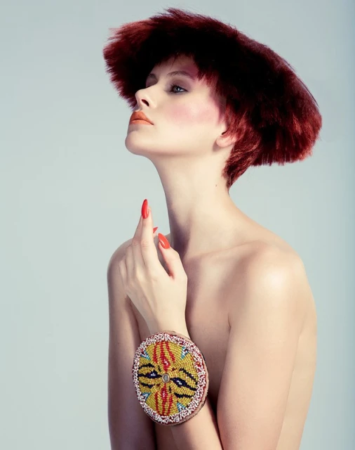 Portrait of a woman with reddish-brown hair and beaded bracelet, looking upward in a studio setting, against a soft grey background.