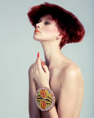 Portrait of a woman with reddish-brown hair and beaded bracelet, looking upward in a studio setting, against a soft grey background.
