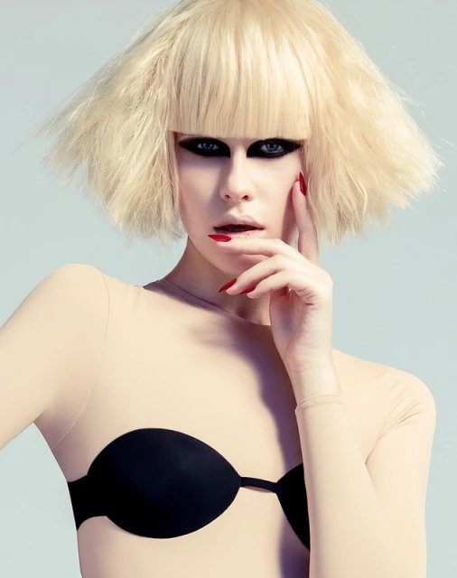 Eye-level shot of a model with a blonde bob haircut, wearing a nude long-sleeved top and black bra, against a light blue backdrop.