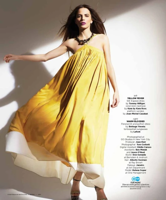 Elegant woman poses in flowing yellow dress with white trim, accessorized with necklace and sandals, against a bright, minimalist background.