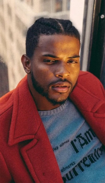 Portrait of a young Black man in a red coat, posing outdoors with natural light, showcasing urban fashion and style against a city backdrop.