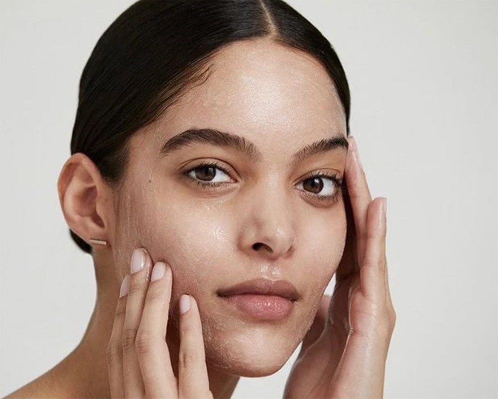 Close-up shot of a woman with dark hair and light skin, applying skincare to her face with her fingers against a neutral background.