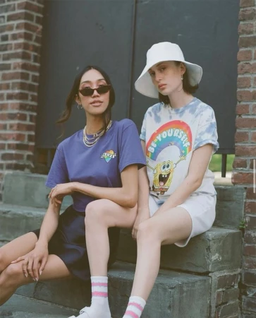 Two young women sit on steps in casual streetwear fashion. One wears a Spongebob graphic tee, the other a violet t-shirt and sunglasses.