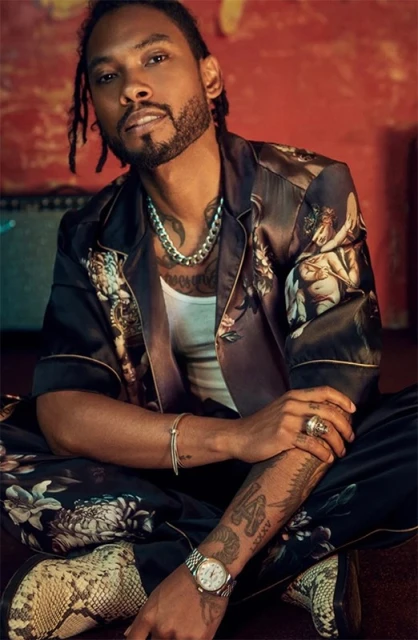 Portrait of a stylish Black man with dreadlocks wearing a printed silk shirt and snake skin boots, sitting indoors against a red wall.