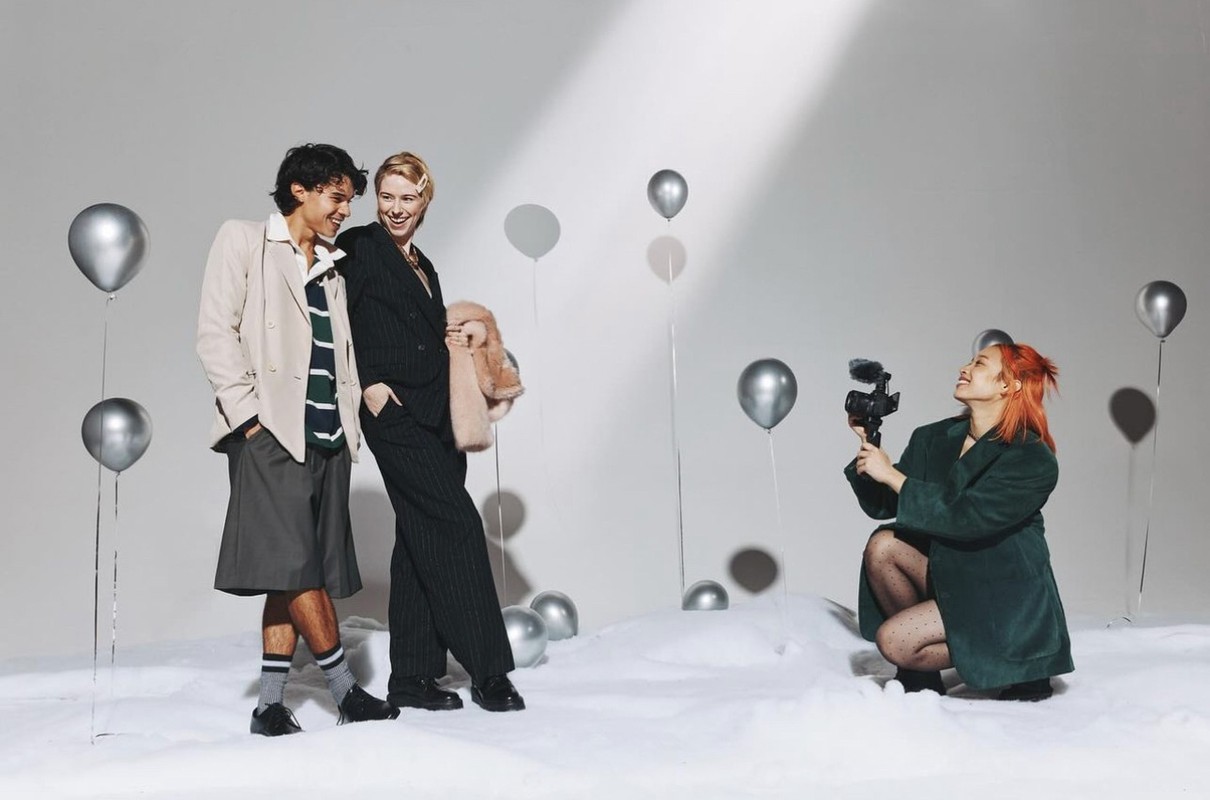 Two people pose near silver balloons, while a person with red hair films with a video camera in a bright photo studio setting.