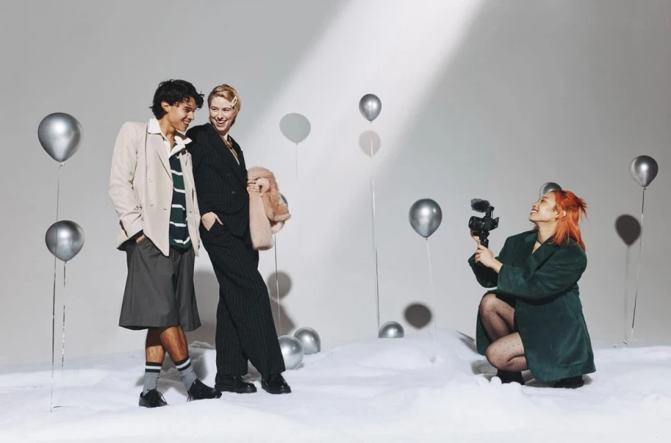Two people pose near silver balloons, while a person with red hair films with a video camera in a bright photo studio setting.