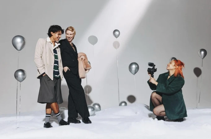 Two people pose near silver balloons, while a person with red hair films with a video camera in a bright photo studio setting.