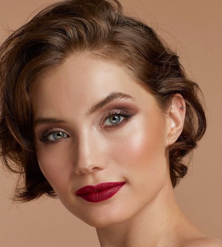 Close-up portrait of a woman with auburn hair, fair skin, dramatic eye makeup, and dark red lipstick against a warm, neutral backdrop.