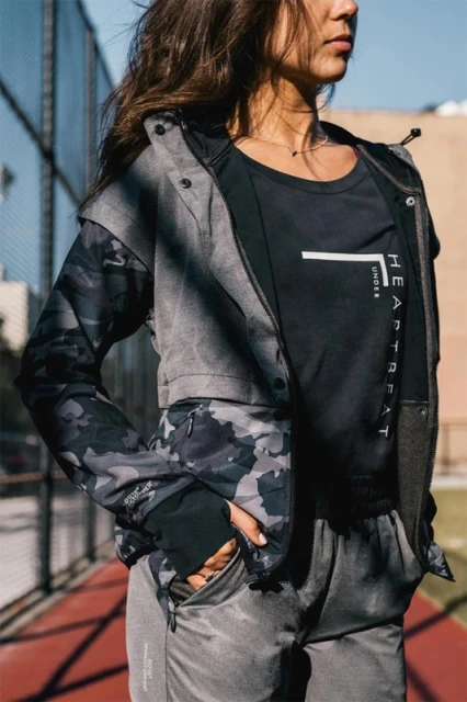 Woman in gray athletic wear and camo jacket poses on an outdoor basketball court in the sunlight with an urban environment in the background.