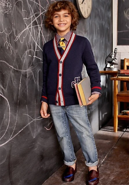 A young boy with curly hair wearing a preppy outfit, including a cardigan, shirt, tie, jeans, and loafers, stands next to a blackboard.