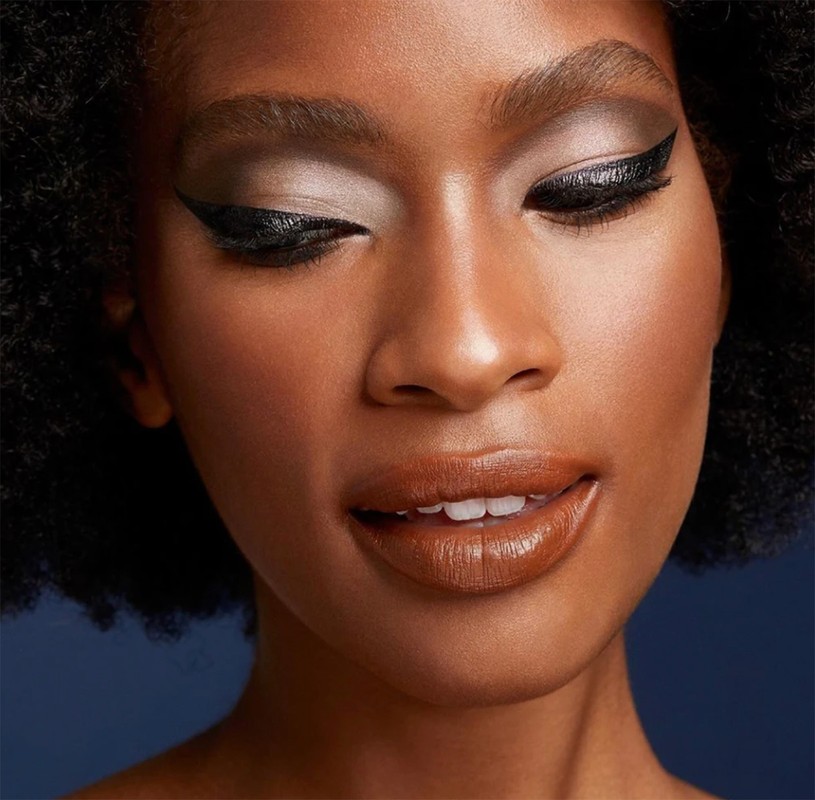 Close-up of a woman with makeup: glossy brown lipstick, defined eyebrows, and black winged eyeliner against a navy blue background.